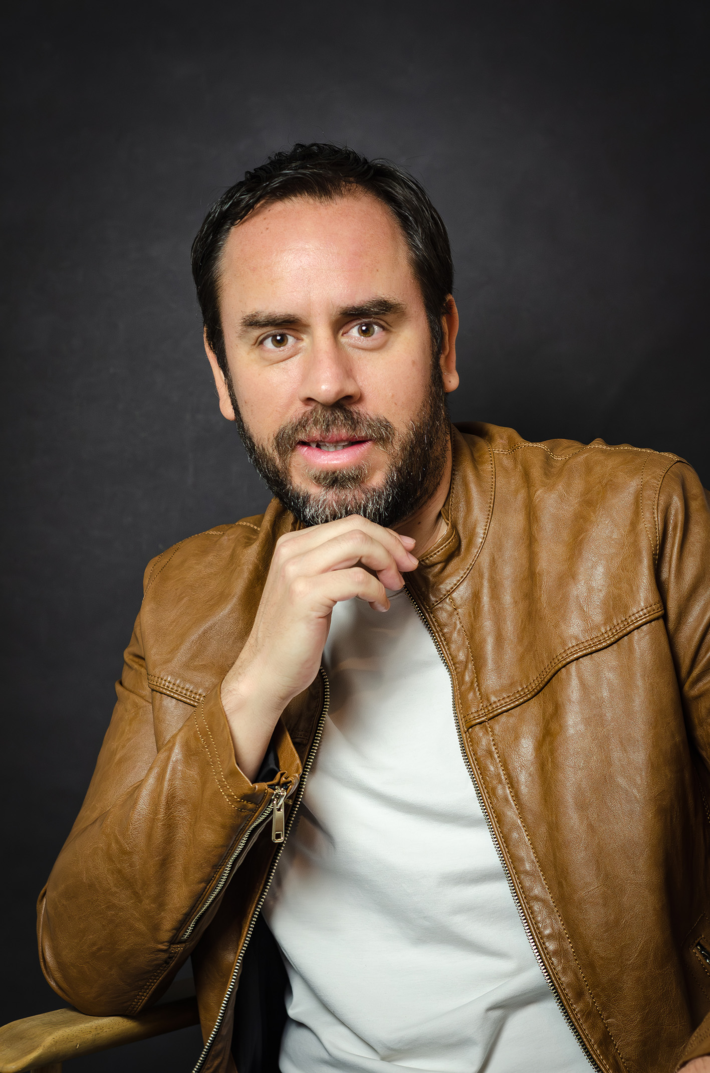 Man with a beard wearing a brown leather jacket and white shirt, posing with a thoughtful expression.